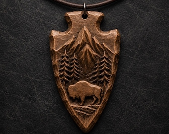 May include: A brown wooden pendant shaped like an arrowhead, suspended from a dark brown cord. The surface features a detailed relief carving of a mountain range, tall pine trees, and a bison standing in the foreground on textured ground.