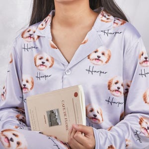 Puede incluir: Conjunto de pijama lila claro con un patrón repetido de la cara de un perro y el nombre "Harper". La parte superior del pijama tiene una botonadura y un cuello. La persona sostiene un libro con el texto "Cafe de Flore".