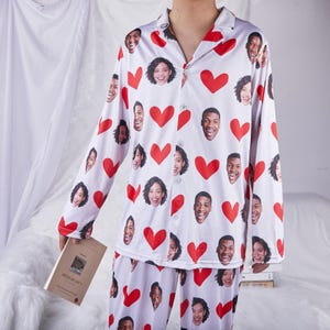 Puede incluir: Conjunto de pijama blanco con un patrón repetido de caras sonrientes y corazones rojos. La parte superior de manga larga tiene una parte delantera abotonada y un cuello. Los pantalones a juego completan el conjunto. La persona sostiene un libro.