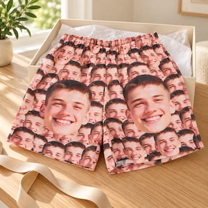 May include: A pair of lounge shorts featuring an all-over repeating pattern of a smiling person's face. The shorts have an elasticized waistband and are presented inside a light-colored gift box lined with white tissue paper on a wooden surface.