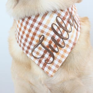 May include: A dog wearing a brown and white checkered bandana with the name "Sully" embroidered in a cursive font. The bandana is a triangle shape and is around the dog's neck.