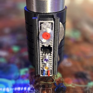 Op de afbeelding: Een zwart en zilver cilindrisch apparaat met een rode knop en verschillende kleinere gekleurde lampjes. Het apparaat heeft een zwarte bovenkant en een zilveren band. Het apparaat is ongeveer 10 cm hoog.