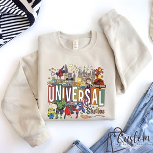 May include: A beige crewneck sweatshirt featuring a colorful graphic of Universal Studios characters and logos. The design includes characters from Jurassic Park, Marvel, and Minions, with the words "UNIVERSAL Studios" in large letters.