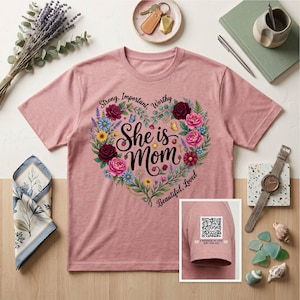 May include: A dusty rose t-shirt with a floral heart design that reads "She is Mom." The heart is filled with roses, daisies, and other flowers. The sleeve has a QR code and the text "A message of love just for you."