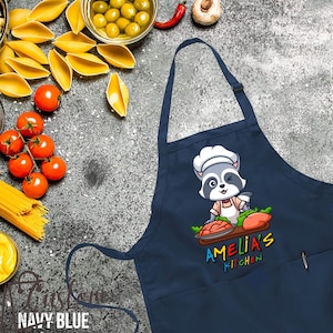 May include: Navy blue kitchen apron with a pocket. A cartoon raccoon chef is holding a plate of food with the text "Amelia's Kitchen".