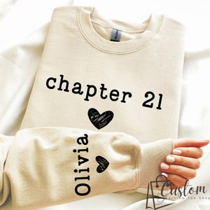May include: Cream-colored sweatshirt with the text "chapter 21" and a black heart graphic. The sleeve features the name "Olivia" in vertical text with two small black heart graphics. A custom fashion tee shop logo is in the bottom right corner.