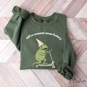 May include: Green sweatshirt with the text "Off to commit mom foolery." The graphic features a green frog wearing a wizard hat and riding a scooter. The sweatshirt has long sleeves and a crew neck.