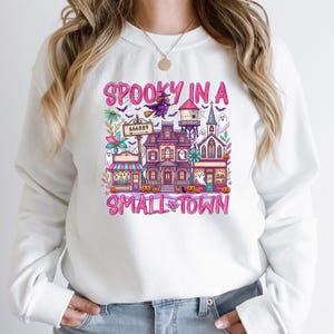 May include: A white long-sleeve sweatshirt featuring a colorful graphic print with the text "SPOOKY IN A SMALL TOWN". The design includes a haunted house, a witch on a broomstick, ghosts, pumpkins, and a signpost labeled "666889" in pink lettering.