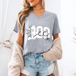 May include: A heather gray V-neck t-shirt featuring the white text BOO with a black spiderweb graphic integrated into the letter B and a small black spider illustration hanging from the letter O, paired with light blue distressed denim shorts.