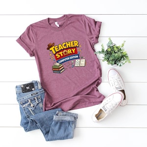 May include: A heathered maroon short-sleeve t-shirt featuring a graphic print that reads "TEACHER STORY COMPUTER EDITION" in yellow and white letters, accompanied by illustrations of a brown hat, stacked books, a computer mouse, and a notebook.