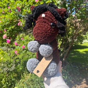 May include: A handmade crochet horse plushie in shades of brown, black, and gray. The horse has a dark brown body, black mane, and gray pom-pom feet. A small tag is attached. The plushie is held up against a background of green foliage and pink flowers.
