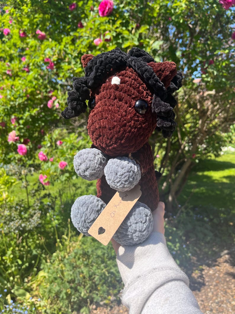 May include: A handmade crochet horse plushie in shades of brown, black, and gray. The horse has a dark brown body, black mane, and gray pom-pom feet. A small tag is attached. The plushie is held up against a background of green foliage and pink flowers.