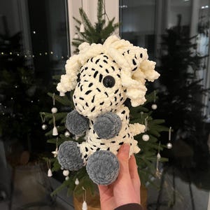 May include: A handmade, plush toy in the shape of a sheep. The sheep is primarily cream-colored with black spots and gray pom-pom feet. It has a large black button eye. The toy is held in front of a Christmas tree.