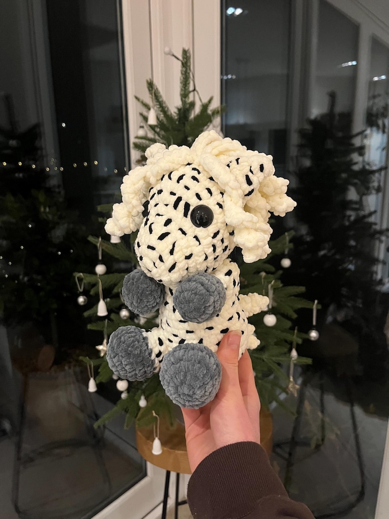 May include: A handmade, plush toy in the shape of a sheep. The sheep is primarily cream-colored with black spots and gray pom-pom feet. It has a large black button eye. The toy is held in front of a Christmas tree.