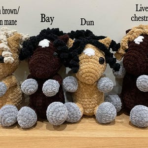May include: Four handmade crochet horse plushies in various colors: warm brown with flaxen mane, bay, dun, and liver chestnut. Each has a white blaze, black eyes, and gray paws. The plushies are sitting on a wooden surface.