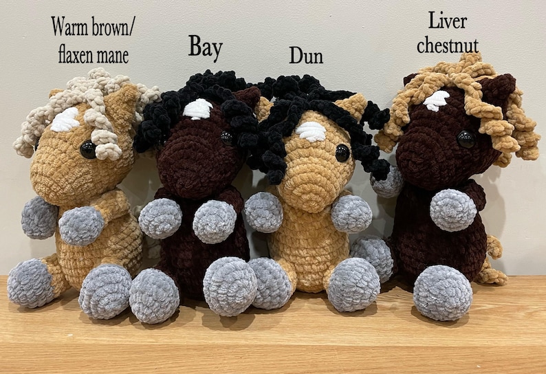 May include: Four handmade crochet horse plushies in various colors: warm brown with flaxen mane, bay, dun, and liver chestnut. Each has a white blaze, black eyes, and gray paws. The plushies are sitting on a wooden surface.