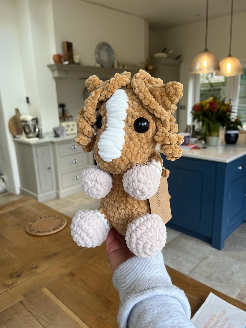 May include: A handmade crochet horse plushie in shades of brown and cream. The horse has a white stripe down its face, large black eyes, and fluffy, rounded features. The plushie is held in a person's hand.
