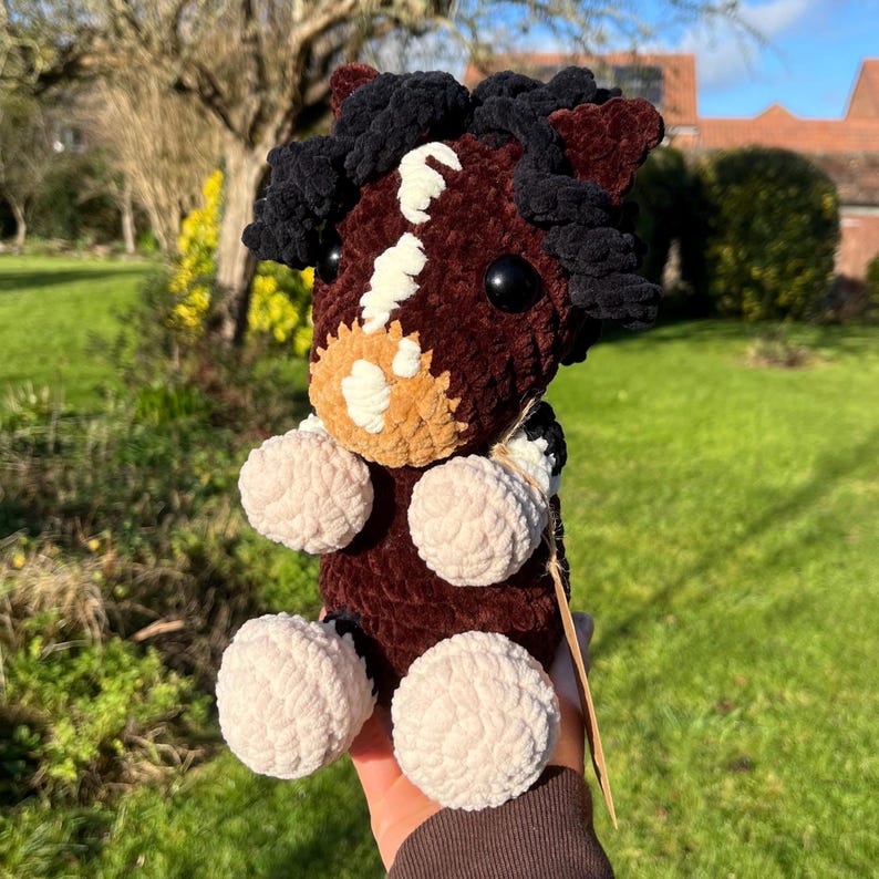 May include: A handmade plush horse toy in shades of brown, black, and cream. The horse features a detailed face with a white blaze, black eyes, and fluffy, cream-colored legs. The toy is made of soft, textured yarn.