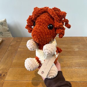 May include: A handmade, plush crochet doll in shades of orange, cream, and beige. The doll has large black eyes and curly orange hair. It is wearing a cream and orange striped sweater. A small tag with the name "George" is attached.
