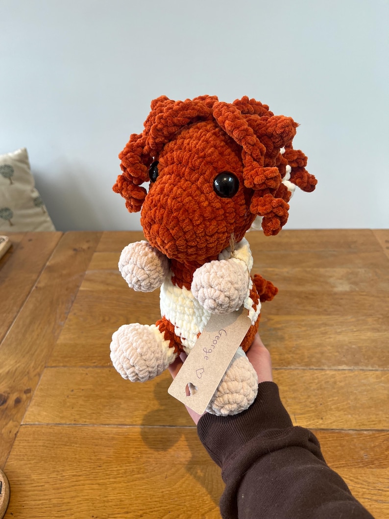 May include: A handmade, plush crochet doll in shades of orange, cream, and beige. The doll has large black eyes and curly orange hair. It is wearing a cream and orange striped sweater. A small tag with the name "George" is attached.