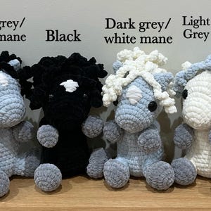 May include: Four handmade crochet horse plushies in various colors. The horses are light grey, black, and dark grey, with manes in black, white, and grey. The text "Dark grey/black mane", "Black", "Dark grey/white mane", and "Light grey/Grey mane" is visible.