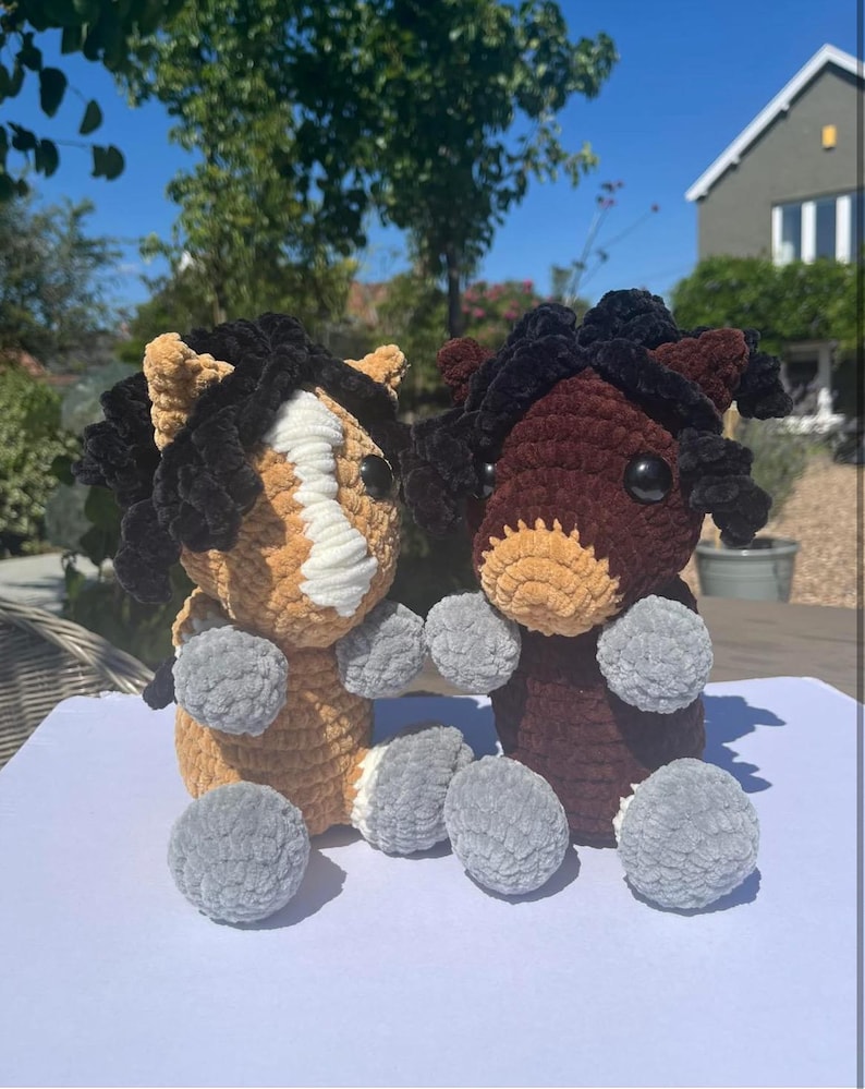 May include: Two handmade crochet horse plushies. One is tan and white, the other is brown. Both have black manes and tails, and gray pom-pom feet. The plushies are sitting on a white surface.