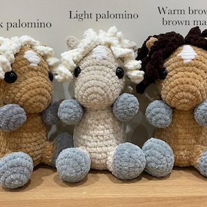 May include: Three handmade crochet horse plushies in varying shades of brown and cream, each with a fluffy mane and tail. The plushies have gray ball-shaped feet and are displayed on a wooden surface.
