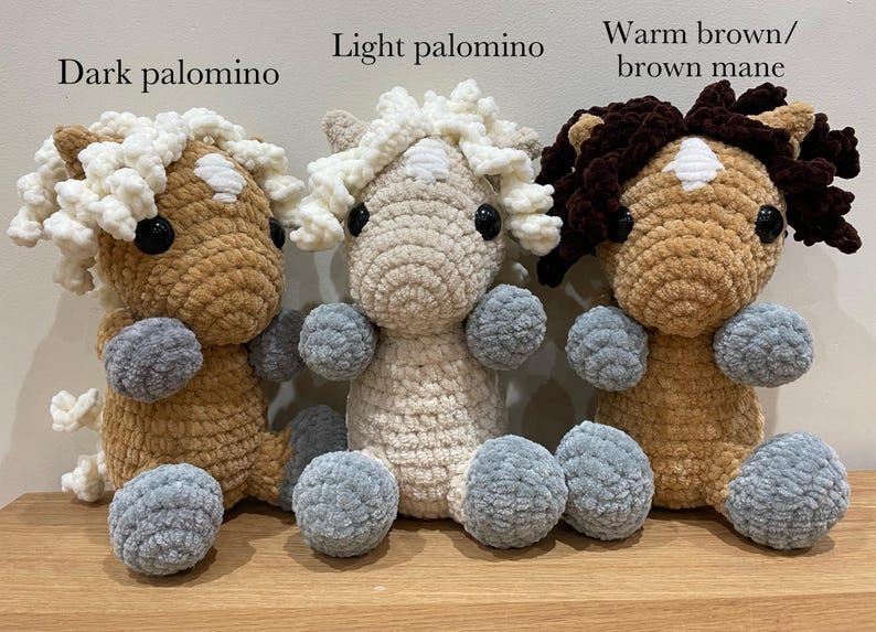 May include: Three handmade crochet horse plushies in varying shades of brown and cream, each with a fluffy mane and tail. The plushies have gray ball-shaped feet and are displayed on a wooden surface.