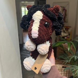 May include: A handmade crochet horse plushie in shades of brown, black, and white. The horse features a fluffy mane and tail, with a white stripe down its face. The plushie has a tag with a heart symbol.