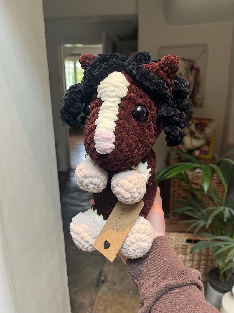 May include: A handmade crochet horse plushie in shades of brown, black, and white. The horse features a fluffy mane and tail, with a white stripe down its face. The plushie has a tag with a heart symbol.
