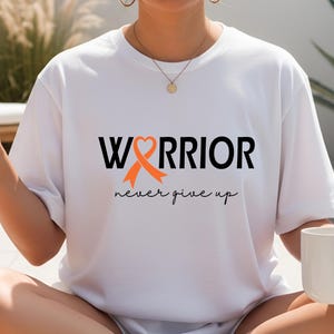 Puede incluir: Una camiseta blanca con el texto negro "WARRIOR", donde una cinta naranja de concienciaci&oacute;n forma la letra "A" con un coraz&oacute;n en la parte superior. Debajo, la frase "never give up" est&aacute; impresa en una fuente cursiva de color negro.