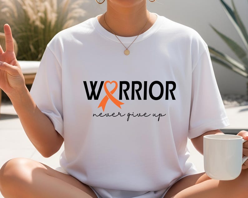 Puede incluir: Una camiseta blanca con el texto negro "WARRIOR", donde una cinta naranja de concienciaci&oacute;n forma la letra "A" con un coraz&oacute;n en la parte superior. Debajo, la frase "never give up" est&aacute; impresa en una fuente cursiva de color negro.