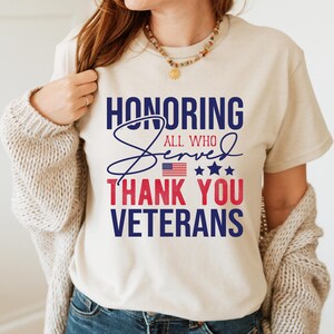 May include: A white t-shirt with a blue and red graphic that says "Honoring All Who Served Thank You Veterans". The graphic includes a small American flag and stars.