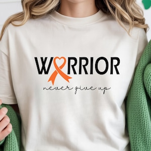 Puede incluir: Una camiseta de manga corta color crema con el texto negro "WARRIOR", donde una cinta de concienciaci&oacute;n naranja forma la letra "A", y el texto cursivo negro "never give up" impreso debajo de la palabra principal en la parte frontal.