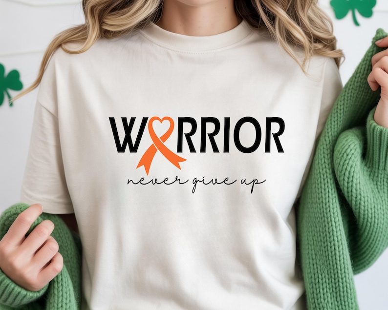 Puede incluir: Una camiseta de manga corta color crema con el texto negro "WARRIOR", donde una cinta de concienciaci&oacute;n naranja forma la letra "A", y el texto cursivo negro "never give up" impreso debajo de la palabra principal en la parte frontal.