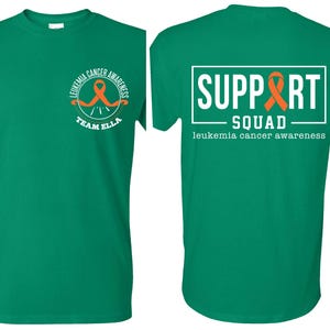 Puede incluir: Una camiseta verde de manga corta con un peque&ntilde;o logotipo circular en el pecho izquierdo que incluye una cinta naranja y el texto "LEUKEMIA CANCER AWARENESS TEAM ELLA". La parte posterior muestra un gran gr&aacute;fico rectangular blanco con "SUPPORT SQUAD" y una cinta naranja.