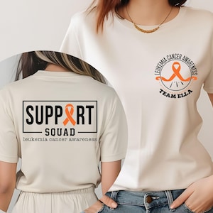 May include: A cream-colored t-shirt with the text "SUPPORT SQUAD" in a black box with an orange ribbon. The shirt also features the words "leukemia cancer awareness" below the box. On the front, a circular design reads "LEUKEMIA CANCER AWARENESS" and "TEAM ELLA".