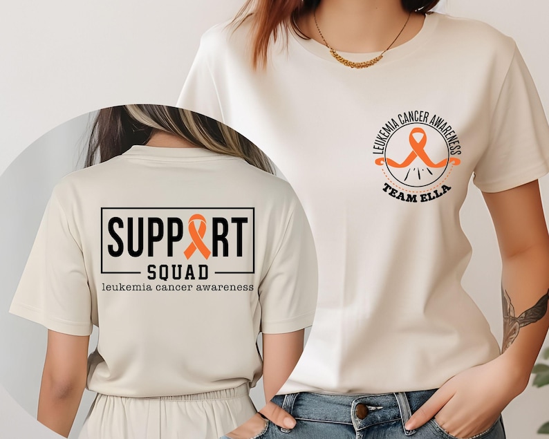Puede incluir: Una camiseta color crema con un dise&ntilde;o de concienciaci&oacute;n sobre la leucemia. El pecho muestra una cinta naranja con el texto LEUKEMIA CANCER AWARENESS TEAM ELLA. La espalda presenta un recuadro con SUPPORT SQUAD y leukemia cancer awareness.