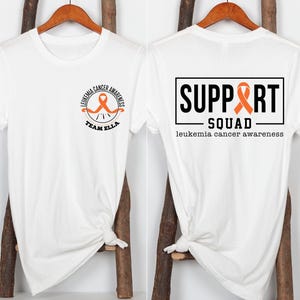 Puede incluir: Una camiseta blanca con un peque&ntilde;o logotipo circular en la parte delantera con una cinta naranja y el texto LEUKEMIA CANCER AWARENESS TEAM ELLA. La parte trasera muestra un gr&aacute;fico rectangular grande con el texto SUPPORT SQUAD y leukemia cancer awareness.