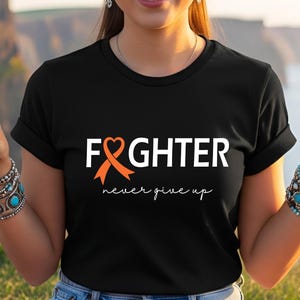 Puede incluir: Una camiseta negra de manga corta con el texto blanco FIGHTER, donde una cinta de concienciaci&oacute;n naranja forma la letra I. Debajo del texto principal, la frase never give up est&aacute; impresa en una fuente cursiva blanca en el centro del pecho.