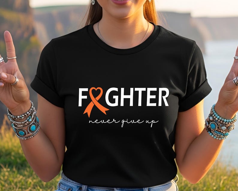 Puede incluir: Una camiseta negra de manga corta con el texto blanco FIGHTER, donde una cinta de concienciaci&oacute;n naranja forma la letra I. Debajo del texto principal, la frase never give up est&aacute; impresa en una fuente cursiva blanca en el centro del pecho.