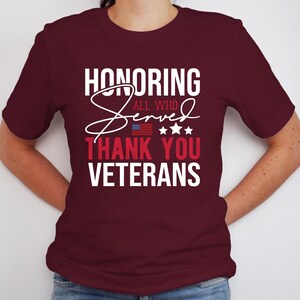 May include: A maroon t-shirt with white text that reads "Honoring All Who Served Thank You Veterans". The text is in a cursive font with a small American flag graphic above the word "Thank".