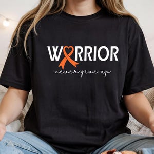 Puede incluir: Una camiseta negra de manga corta con el texto blanco "WARRIOR", donde la letra "A" es reemplazada por un lazo de concienciaci&oacute;n naranja con forma de coraz&oacute;n. Debajo, la frase "never give up" est&aacute; impresa en una fuente cursiva blanca.