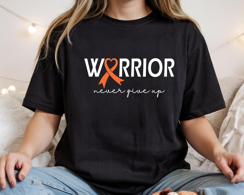 Puede incluir: Una camiseta negra de manga corta con el texto blanco "WARRIOR", donde la letra "A" es reemplazada por un lazo de concienciaci&oacute;n naranja con forma de coraz&oacute;n. Debajo, la frase "never give up" est&aacute; impresa en una fuente cursiva blanca.