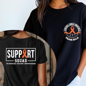 Puede incluir: Una camiseta negra con dos dise&ntilde;os gr&aacute;ficos distintos. El pecho muestra una cinta naranja con el texto LEUKEMIA CANCER AWARENESS TEAM ELLA. La parte posterior presenta un marco rectangular blanco con el texto SUPPORT SQUAD.