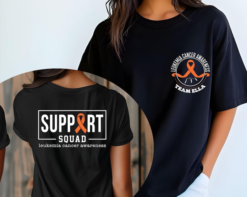 Puede incluir: Una camiseta negra con dos dise&ntilde;os gr&aacute;ficos distintos. El pecho muestra una cinta naranja con el texto LEUKEMIA CANCER AWARENESS TEAM ELLA. La parte posterior presenta un marco rectangular blanco con el texto SUPPORT SQUAD.