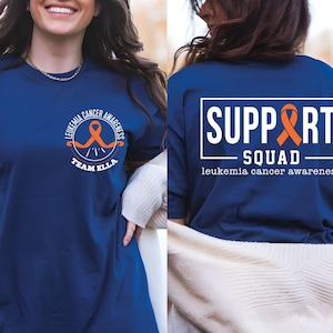 Puede incluir: Una camiseta azul marino con un gr&aacute;fico de cinta naranja circular en el pecho izquierdo con el texto LEUKEMIA CANCER AWARENESS TEAM ELLA. La parte posterior muestra un gran marco rectangular blanco con el texto SUPPORT SQUAD y una cinta.
