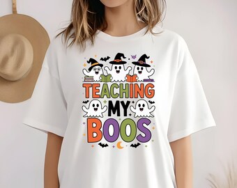 May include: White t-shirt with a Halloween-themed graphic. The design features three cartoon ghosts wearing witch hats and reading books above the text "TEACHING MY BOOS". Additional ghosts, bats, and a crescent moon are also included in the design.