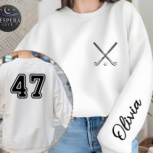 May include: White sweatshirt with black accents. Features a crossed hockey stick and ball graphic on the chest, the name "Olivia" on the sleeve, and the number "47" on the back. The sweatshirt has a crew neck and a relaxed fit.