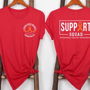 Puede incluir: Una camiseta roja con un gr&aacute;fico frontal que muestra una cinta naranja y el texto "LEUKEMIA CANCER AWARENESS TEAM ELLA". El gr&aacute;fico trasero muestra un marco rectangular blanco con el texto "SUPPORT SQUAD" y "leukemia cancer awareness".
